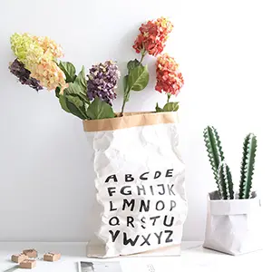 Colorful artificial flowers in a paper vase with alphabet design, next to a potted cactus.